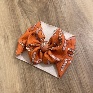 Könnte beinhalten: Ein Baby-Stirnband mit einer großen Schleife mit einem sich wiederholenden Muster aus Zuckerstangen auf einem strukturierten orangefarbenen Hintergrund. Das Stirnband ist weiß und die Schleife hat rosa und weiße Zuckerstangen-Designs.