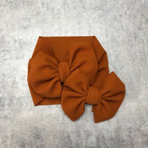 May include: Two brown fabric headbands with large bows. The larger headband has a single bow, while the smaller headband has a smaller bow.
