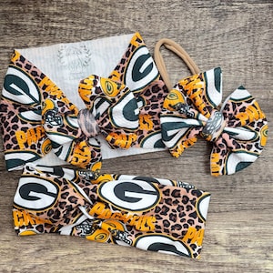 May include: A set of Green Bay Packers themed hair accessories, including a headband and several bows. The items feature a leopard print pattern with the team's logo in green, yellow, and white. The accessories are displayed on a wooden surface.