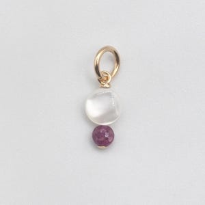 Può includere: Un pendente dorato con un anello, caratterizzato da una pietra trasparente e arrotondata e una gemma rosa scuro sfaccettata più piccola. Il pendente è posto su uno sfondo bianco.