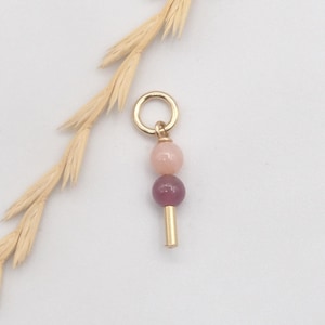 Peut inclure: Un pendentif en métal doré composé d'un anneau circulaire en haut, suivi d'une perle sphérique rose clair, d'une perle sphérique magenta foncé et d'un court cylindre en métal doré lisse à la base, disposés en ligne verticale.