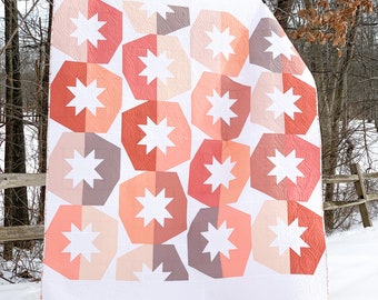 Puede incluir: Una colcha con un patrón geométrico de estrellas blancas sobre un fondo rosa, naranja y marrón. La colcha está colgada de un tendedero en un paisaje invernal nevado.