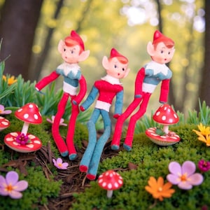 May include: Three vintage-style elf figurines with red and blue outfits and pointed hats stand among artificial mushrooms and flowers. The elves have long, thin legs and arms. The scene is set on a bed of green moss.