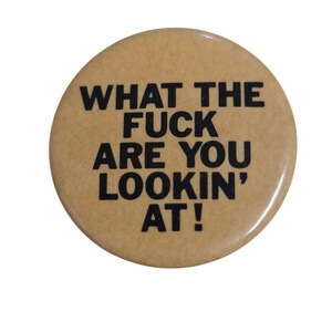 Peut inclure: Un bouton rond beige avec l'inscription "WHAT THE FUCK ARE YOU LOOKIN' AT !" en lettres noires grasses. Le bouton a une finition brillante et un design simple et direct.