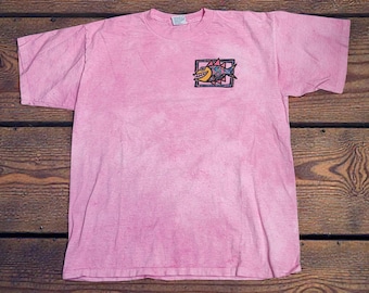 May include: Pink t-shirt with a small, colourful fish graphic in a square frame on the chest. The shirt has a faded appearance and a crew neck. The fish graphic features a smiling face and a mix of colours.