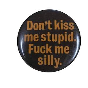 Peut inclure: Un bouton rond noir avec le texte "Don't kiss me stupid. Fuck me silly." en orange. Le bouton a une finition brillante et un design simple et audacieux.