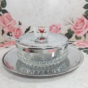 May include: A silver serving dish with a clear, faceted glass bowl and lid, on a matching tray. The lid has a clear handle with a red floral design. A small spoon rests on the rim. The background has a floral pattern.