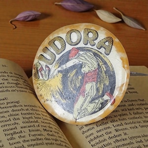 May include: Vintage round button with the word "NUDORA" in bold letters. The button features an illustration of a figure in motion, wearing a red sash and head covering, set against a dark background. The button is placed on an open book.