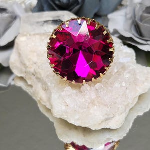 May include: A round, faceted fuchsia-colored gemstone ring set in a gold-toned metal prong setting. The ring is positioned on top of a clear, jagged quartz crystal cluster, showcasing the vibrant pink stone and its reflective, geometric cut.