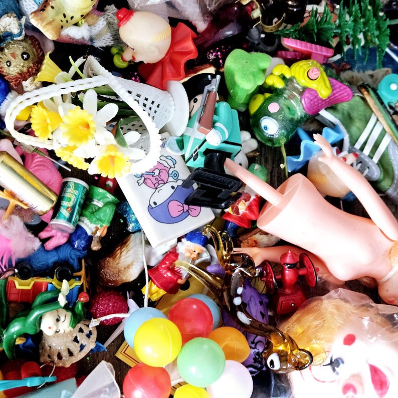 May include: A collection of colorful plastic toys, including a doll, a toy soldier, a toy fish, a toy flower, and a toy balloon. The toys are arranged in a pile and some are partially obscured by other toys.