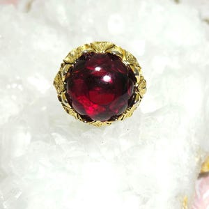 May include: A gold ring featuring a large, round, deep red gemstone. The gemstone is encircled by a decorative gold setting with a leaf design. The ring is set against a white, textured backdrop.