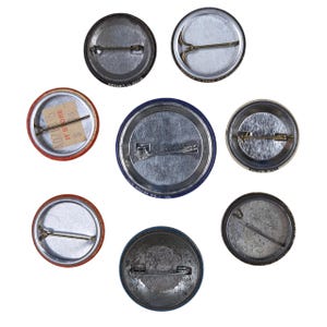Peut inclure: Ensemble de huit badges ronds, vus de dos, avec des fermoirs et des &eacute;pingles en m&eacute;tal. Les boutons pr&eacute;sentent des couleurs et des motifs vari&eacute;s, certains montrant des signes d'usure. Un bouton a une &eacute;tiquette de prix attach&eacute;e.
