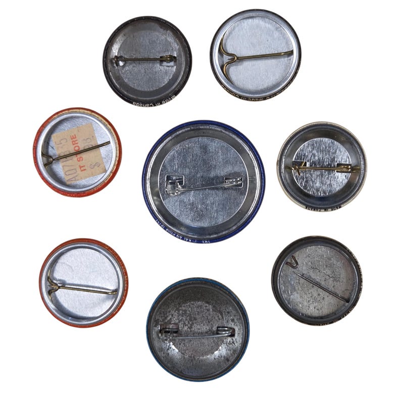Peut inclure: Ensemble de huit badges ronds, vus de dos, avec des fermoirs et des &eacute;pingles en m&eacute;tal. Les boutons pr&eacute;sentent des couleurs et des motifs vari&eacute;s, certains montrant des signes d'usure. Un bouton a une &eacute;tiquette de prix attach&eacute;e.