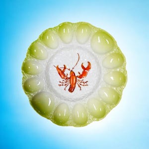 May include: A decorative serving plate with a central design of a red lobster. The plate has a light green, scalloped edge with 12 egg-shaped indentations. The center is white with a textured pattern.
