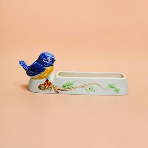 May include: A ceramic pen holder featuring a blue and yellow bird perched on one end. The base is off-white and elongated with a hollow centre, decorated with a painted brown branch and small green leaves extending along the front side.