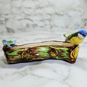 May include: A ceramic log-shaped planter, featuring a blue, yellow, and green bird figurine. The planter is painted with brown, green, and yellow details. A small, colorful fish decorates the other end of the planter.