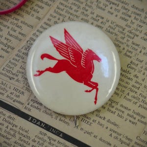 May include: A round, off-white button with a red Pegasus design. The winged horse is depicted mid-leap, with its wings spread. The button has a pin on the back, suggesting it's a wearable accessory. The design is simple and bold.