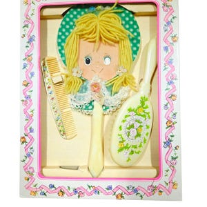 May include: A vintage children's hair care set in a decorative box. The set includes a mirror, brush, and comb, all with a cream-colored finish. The mirror features a cartoon girl design with yellow yarn hair. The box is decorated with pink ribbons and floral patterns.