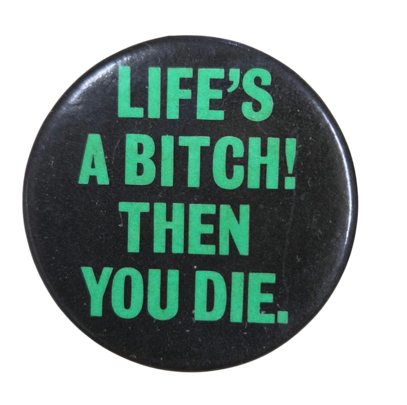Peut inclure: Un bouton rond noir avec l'inscription verte &laquo; LIFE'S A BITCH! THEN YOU DIE &raquo;. Le bouton a une finition brillante et un message simple et direct.