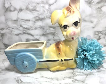 May include: A vintage ceramic planter shaped like a yellow dog pulling a light blue cart. The dog has brown spots and a pink nose. A blue flower sits next to the dog. The planter is likely for small plants or flowers.