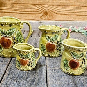 May include: Set of four green ceramic jugs with a fruit and leaf design. The jugs are different sizes and have a handle on one side.