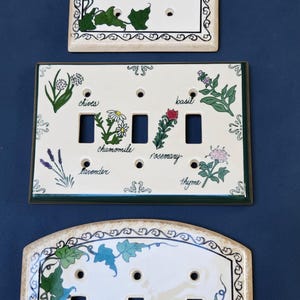 May include: Three ceramic light switch plates. The top and bottom plates feature green ivy vines on a cream background. The middle rectangular plate displays illustrations of chives, basil, chamomile, rosemary, lavender, and thyme with labels.