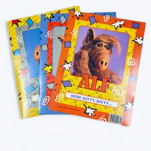 May include: A stack of vintage ALF notebooks. The covers feature the ALF character with the text "ALF" and "HERE KITTY, KITTY...". The notebooks have colorful designs with stars, squares, and dots. The background is white.