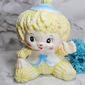 May include: A vintage ceramic figurine of a child with yellow hair and a blue hat. The figurine is wearing a white and blue outfit and has rosy cheeks. The child is in a seated position with one arm raised.