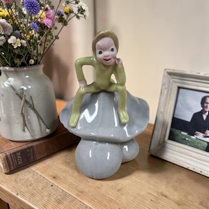 May include: A ceramic figurine of a small, light green elf with a pink face and green eyes, sitting atop a smooth, glossy, light gray mushroom. The elf has a pointed cap and is posed with one hand resting on its chin and the other on its hip.