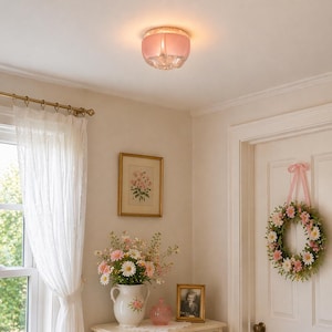 May include: A pink and clear glass ceiling light. The light fitting has a frosted pink glass shade and a clear, faceted glass base. It's mounted on a white ceiling in a room with a floral wreath and framed artwork. The room has a vintage aesthetic.