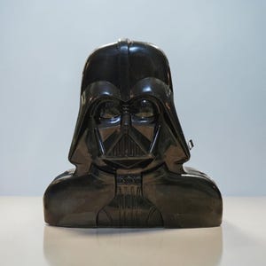 May include: A black Darth Vader bust, a collectible item from the Star Wars franchise. The bust features the character's iconic helmet and mask design, with intricate details and a glossy finish. The bust is a dark, solid color.