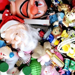 May include: A collection of colorful, whimsical figurines and trinkets, including a Santa Claus, a clown, a teddy bear, and a small bottle of liquor. The figurines are arranged on a white surface with a variety of other decorative items.