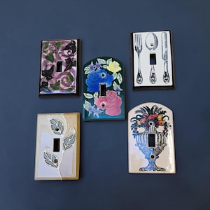 May include: Five rectangular and arched light switch plates featuring various designs: a purple ivy pattern, a floral arrangement with blue and pink roses, a black and white cutlery set, a beige plate with stylised leaves, and a fruit-filled urn.
