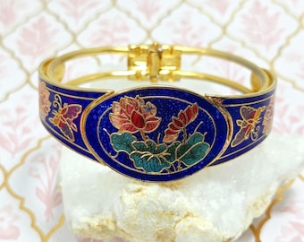 May include: A gold-toned hinged bracelet with a blue enamel band. The bracelet features a floral design with red, green, and blue accents. Butterflies are also depicted on the bracelet. The bracelet is displayed on a white surface.
