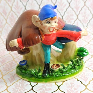 以下が含まれることがあります： 青い帽子、赤いシャツ、ターコイズ色のズボンを身に着けた、気まぐれなエルフの陶器製フィギュア。茶色のキノコのそばに立っています。ベースには緑の草、小さなキノコ、青い花があります。フィギュアは陽気な表情をしています。