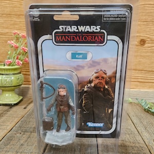 K&ouml;nnte beinhalten: Eine Star Wars The Mandalorian Actionfigur von Kuiil, einem Charakter aus der Serie. Die Figur ist in einer durchsichtigen Plastik-Blisterverpackung mit blau-wei&szlig;em Hintergrund verpackt. Die Figur tr&auml;gt eine braune Lederjacke und eine Schutzbrille. Die Verpackung tr&auml;gt auch ein Kenner-Logo.
