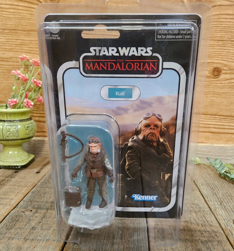 K&ouml;nnte beinhalten: Eine Star Wars The Mandalorian Actionfigur von Kuiil, einem Charakter aus der Serie. Die Figur ist in einer durchsichtigen Plastik-Blisterverpackung mit blau-wei&szlig;em Hintergrund verpackt. Die Figur tr&auml;gt eine braune Lederjacke und eine Schutzbrille. Die Verpackung tr&auml;gt auch ein Kenner-Logo.