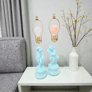 May include: Two light blue table lamps with cherub bases and gold-colored bulb holders. One lamp has a white bulb, the other a pink bulb. A white vase with yellow flowers sits nearby on a white table.