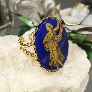May include: A gold-toned ring featuring an oval-shaped deep blue cabochon with a gold-coloured illustration of a figure in flowing robes. The ring band is constructed from a twisted metal wire design, and the cabochon is set within a scalloped edge.