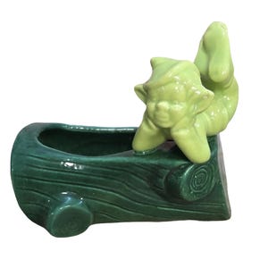May include: A ceramic log-shaped planter with a green elf figure resting on the side. The planter is dark green, and the elf is a lighter shade of green. The planter is designed to hold plants.