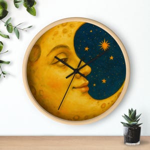 Pode incluir: Relógio de parede redondo com uma face de lua crescente amarela e um céu estrelado azul escuro. O relógio tem uma moldura de madeira clara e ponteiros pretos. O mostrador do relógio retrata uma lua serena.