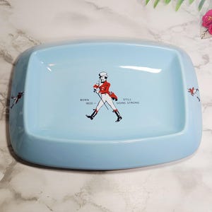 May include: Light blue rectangular ceramic dish with rounded corners. The dish features a graphic of a man in a red coat and black boots, with the text "BORN 1820 - STILL GOING STRONG". Additional figures are on the sides.