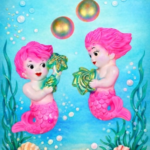 May include: Two pink-haired mermaid figurines holding sea shells, set against a blue background with bubbles and seaweed. The mermaids have pink tails and are holding green and gold shells. Two iridescent orbs float above them.