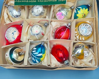 Peut inclure: Une boîte en carton contenant douze décorations de sapin de Noël assorties dans des compartiments individuels. La boîte présente une étiquette verte avec le texte "unbreakable christmas tree". Les ornements varient en forme, incluant des styles ronds et en goutte, avec des finitions métalliques, rouges, bleues et jaunes, certains arborant des motifs floraux peints et des indentations réfléchissantes.