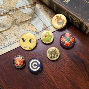 May include: A collection of seven circular pinback buttons featuring various designs, including a cream button with black text V.G. W.S., a green leaf emblem with O.A.F., a purple button with a moose, and a blue button with a white letter C.