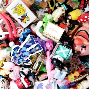 May include: A colorful collection of small toys and trinkets, including a robot, a clown, a frog, a cross, a book, and a strawberry. The toys are arranged in a pile and are mostly made of plastic.