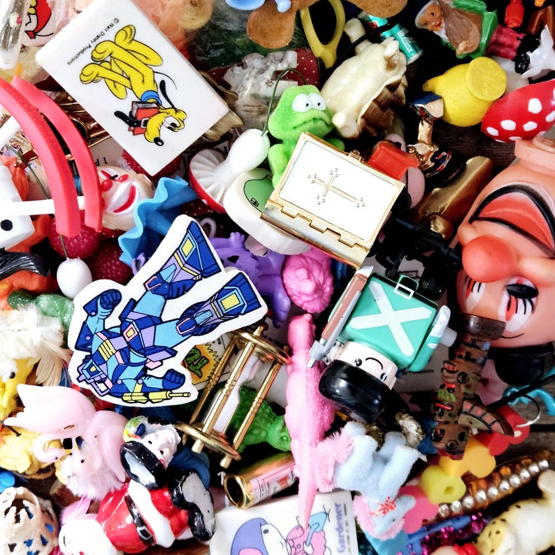 May include: A colorful collection of small toys and trinkets, including a robot, a clown, a frog, a cross, a book, and a strawberry. The toys are arranged in a pile and are mostly made of plastic.