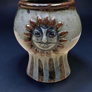 May include: A ceramic chalice featuring a light grey crackle glaze and a brown rim. The body displays a raised, sculpted sun face with brown rays. The base is decorated with vertical brown and grey stripes, creating a distinct contrast.