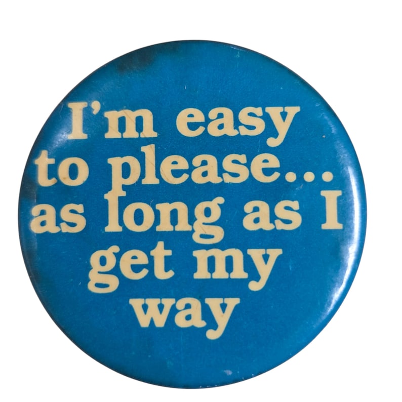 Peut inclure: Un bouton rond bleu avec le texte "I'm easy to please... as long as I get my way" en cr&egrave;me. Le bouton pr&eacute;sente une l&eacute;g&egrave;re usure.