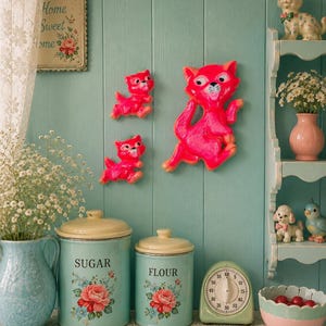 May include: A collection of kitchen decor including two bright pink cat wall plaques, a light blue pitcher, two light blue canisters labelled SUGAR and FLOUR with rose decals, a light green kitchen timer, and a pink bowl containing red cherries.